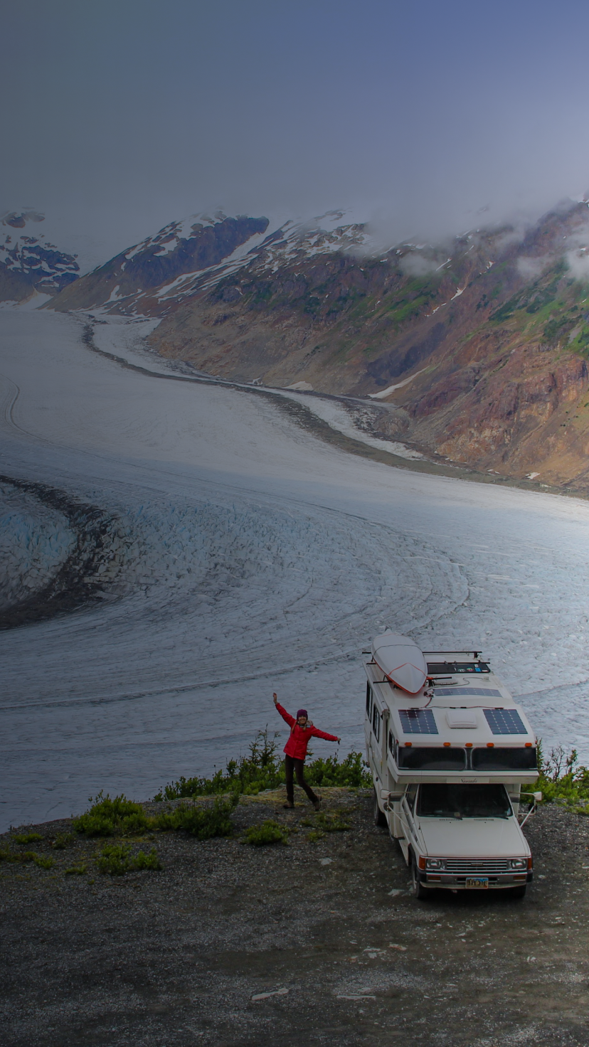 A woman posing next to an RV on a cliff with a snowy glacier below.
